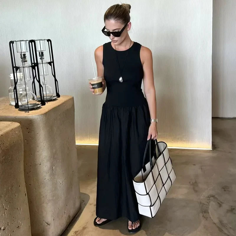Woman in a black dress holding a white tote bag and a drink, standing in a modern indoor setting.